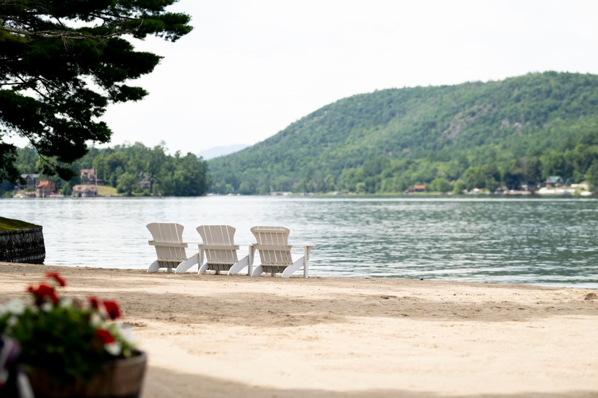 White Adirondack chairs on the sandy beach at Brant Lake, with forested hills across the water